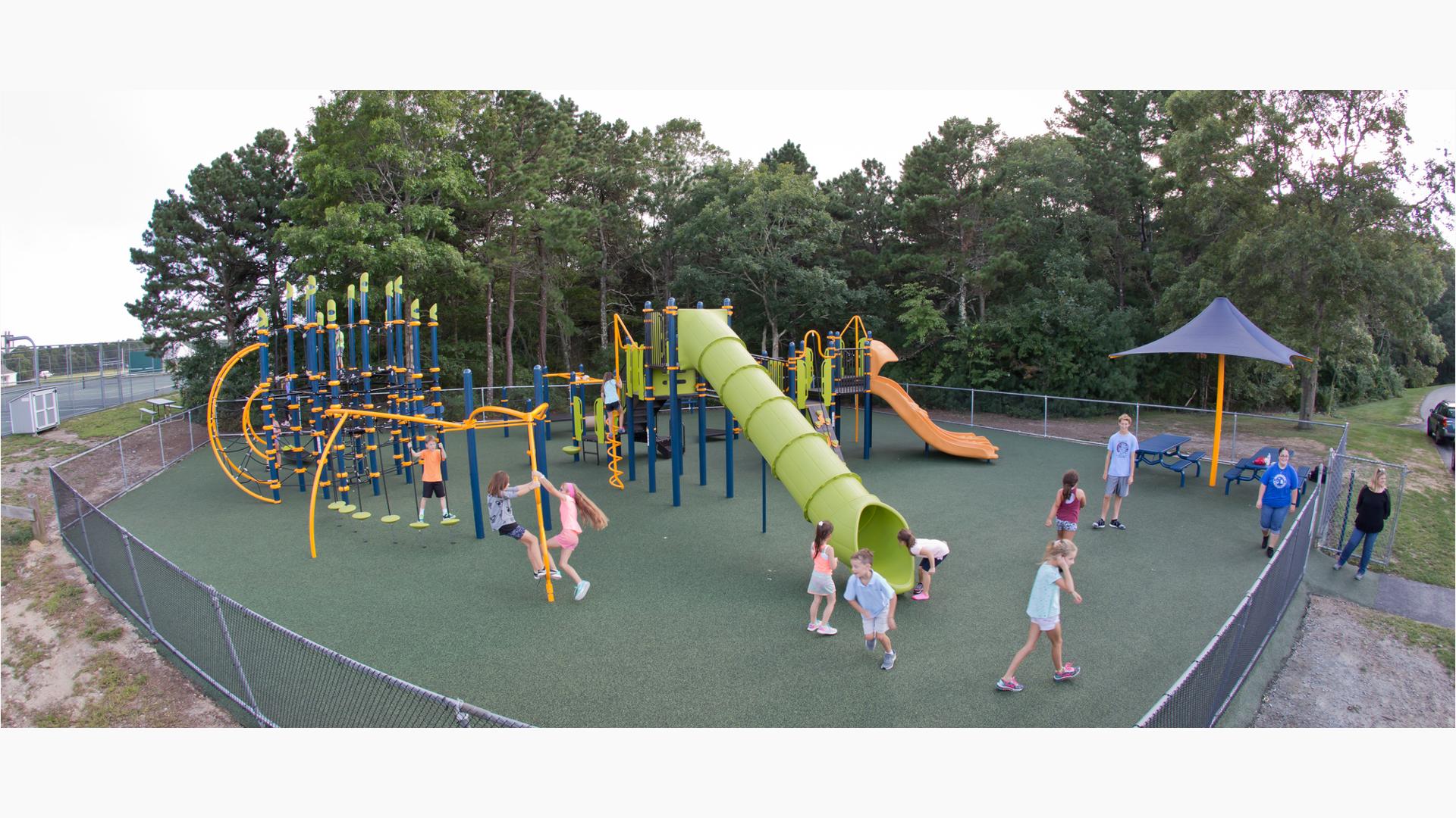 Kids from Quashnet Schoolhaving a good time on their playground. Two girls look up a tunnel slide while a boy leaps out. Two girls play on a Gyro Twister Spinner and a boy plays stands on Disc Challenge.