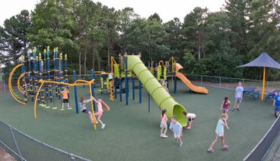 Kids from Quashnet Schoolhaving a good time on their playground. Two girls look up a tunnel slide while a boy leaps out. Two girls play on a Gyro Twister Spinner and a boy plays stands on Disc Challenge.