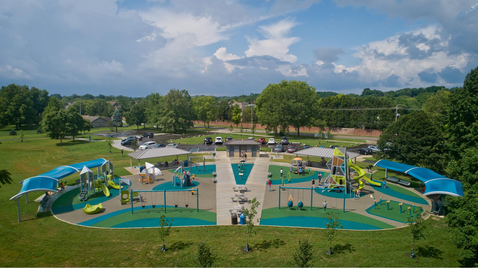 Full elevated view of a large park play area with tower structures surrounded by many other playground activities. Seating areas on either side of the play area have large blue shades overhead. 