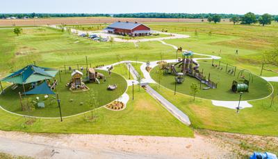 Elevated view of two circular shaped play areas containing nature-inspired play structures. A large open grassy field leads to a large red barn building in the background. 