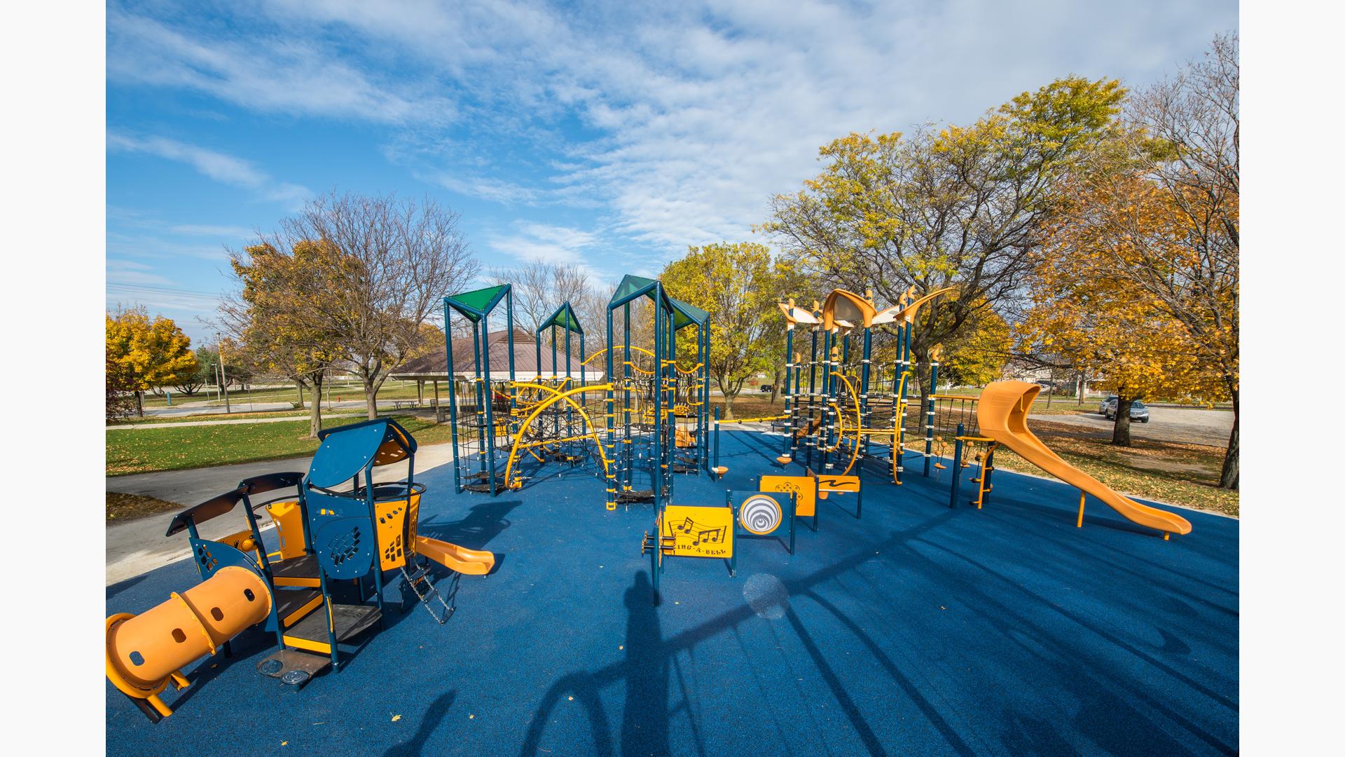 Large playspace that includes tall climbing net structures for older kids and a play house themed playstructure for younger kids. Playspace includes blue surfacing and fall leave colors in the background. 