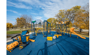 Large playspace that includes tall climbing net structures for older kids and a play house themed playstructure for younger kids. Playspace includes blue surfacing and fall leave colors in the background. 