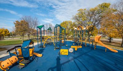 Large playspace that includes tall climbing net structures for older kids and a play house themed playstructure for younger kids. Playspace includes blue surfacing and fall leave colors in the background. 