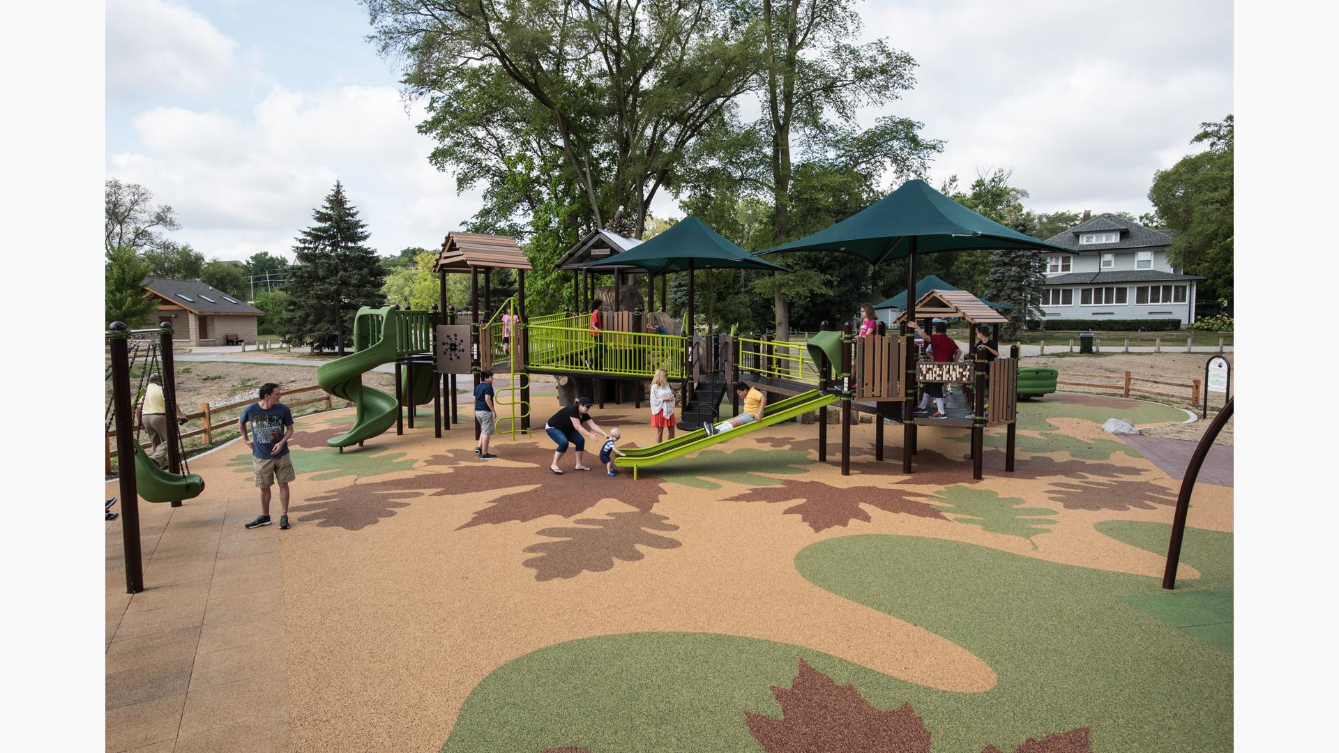 Nature-inspired playground with kids playing and parents joining in. Surfacing includes a leaf design colored in green and brown. 