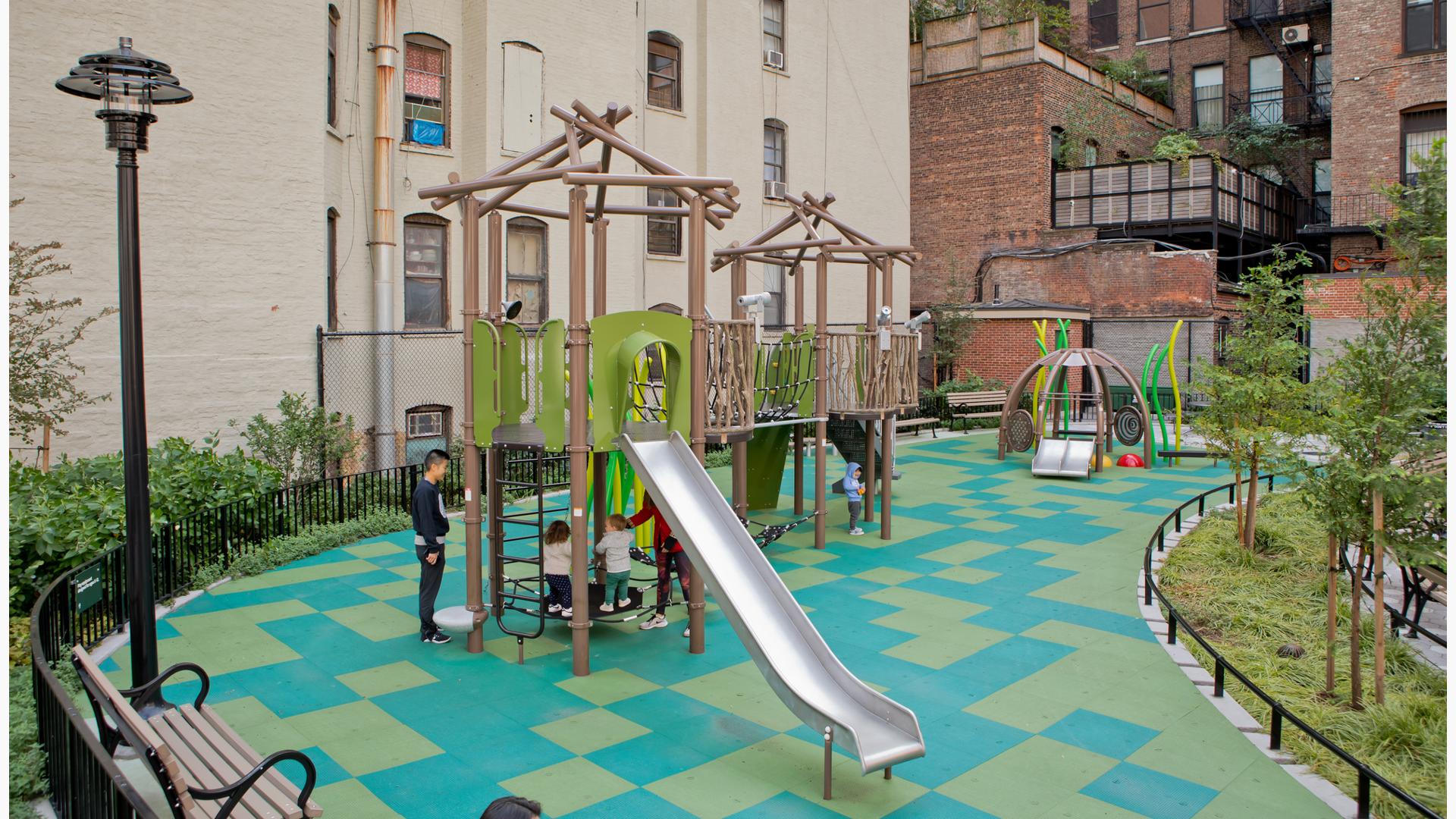 Children play on the multi green square playground surfacing of a nature themed play space amongst surrounding apartment buildings.