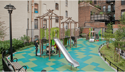 Children play on the multi green square playground surfacing of a nature themed play space amongst surrounding apartment buildings.