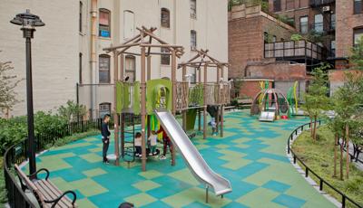 Children play on the multi green square playground surfacing of a nature themed play space amongst surrounding apartment buildings.