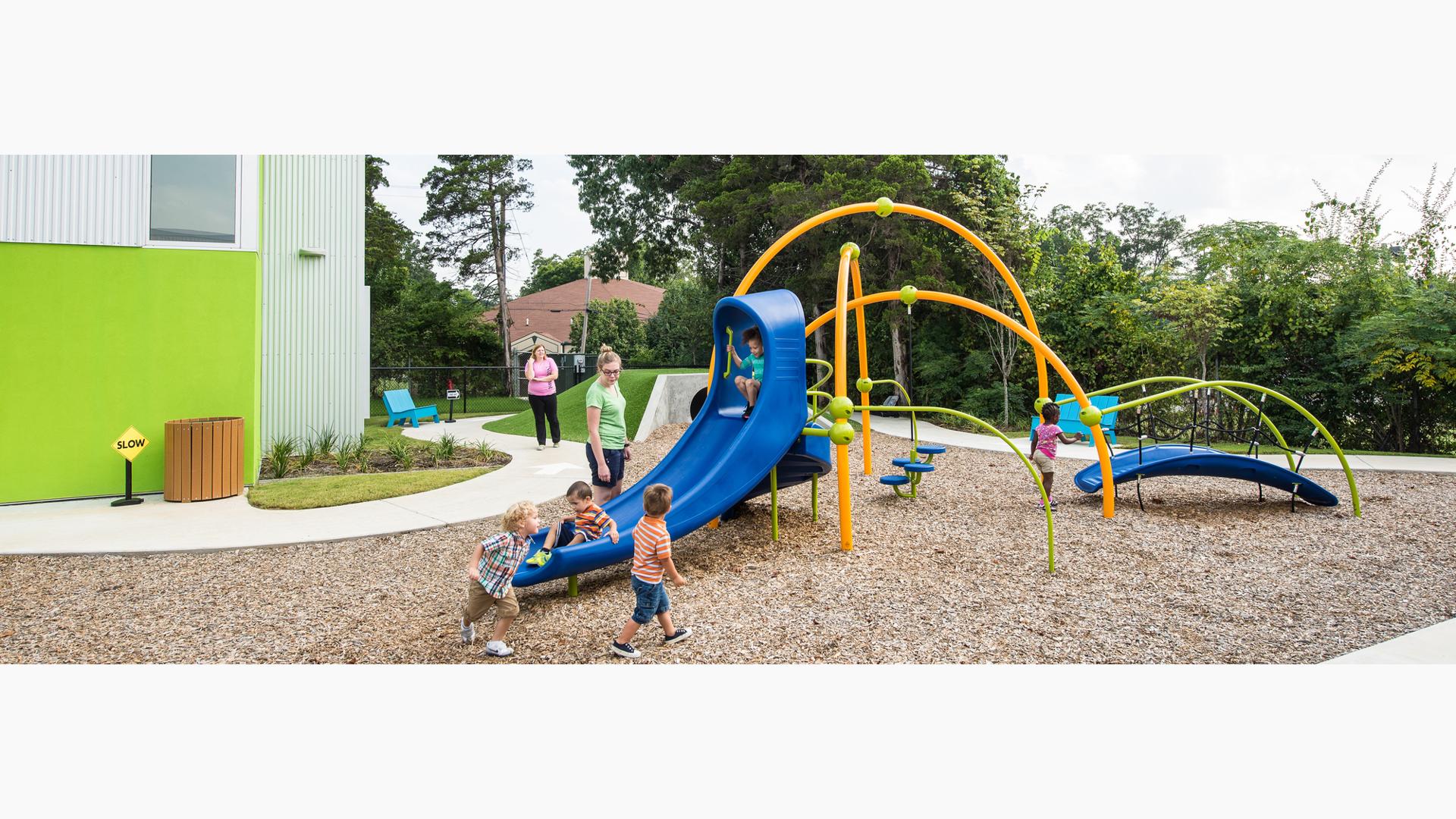 Three orange arched playground posts have a walking bridge, slide, and pod stepper connected to them. Children riding down blue slide as a woman stands nearby watching.