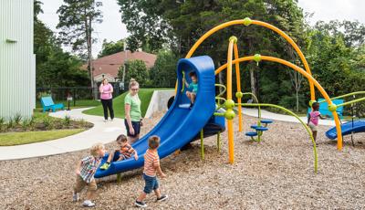 Three orange arched playground posts have a walking bridge, slide, and pod stepper connected to them. Children riding down blue slide as a woman stands nearby watching.