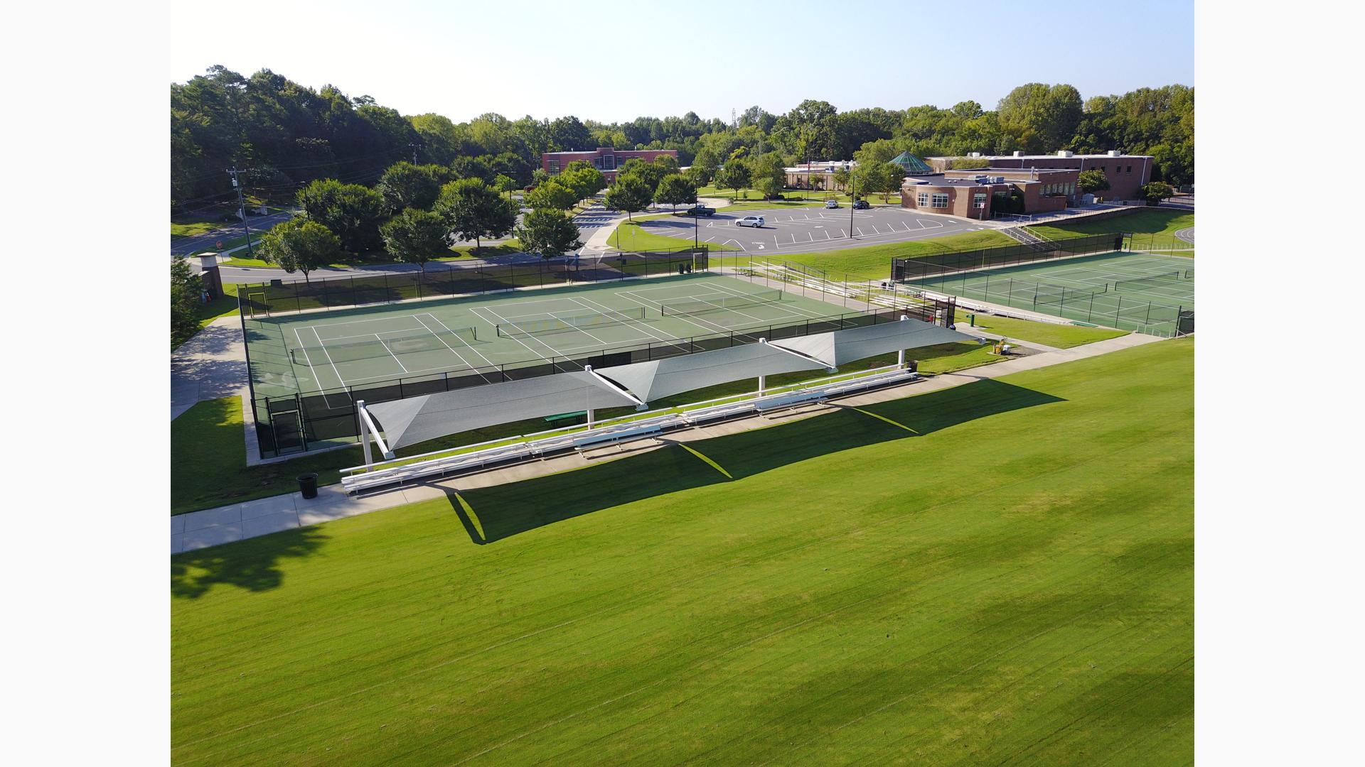 Elevated view of a lush green field with a sidewalk containing rows of bleachers covered by three large rectangular shade systems and a fenced in location behind with three tennis courts. 