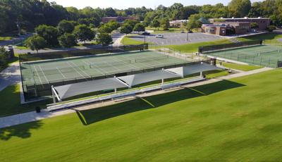 Elevated view of a lush green field with a sidewalk containing rows of bleachers covered by three large rectangular shade systems and a fenced in location behind with three tennis courts. 