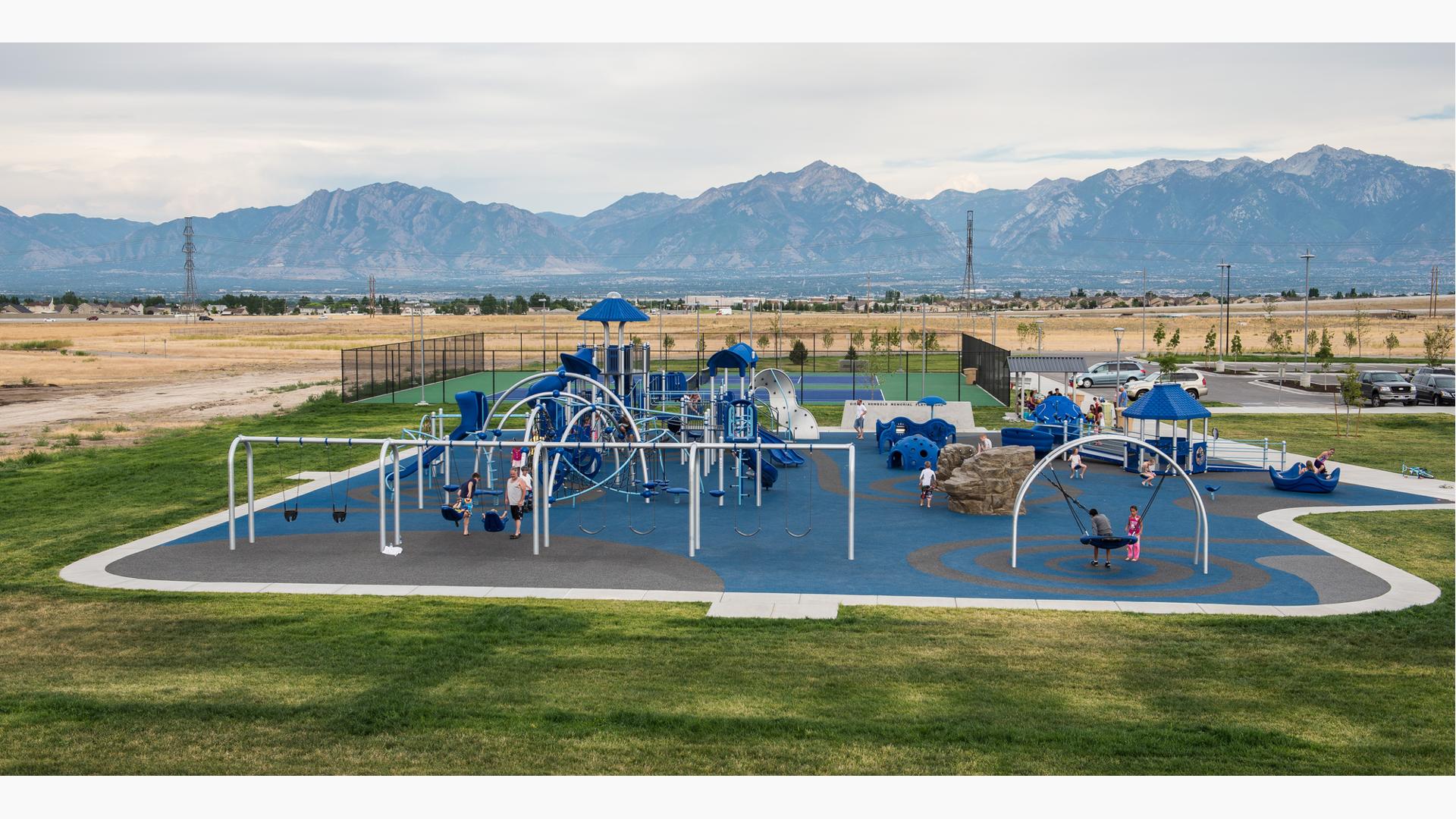 This playground is named for a 6-year-old kidnapping victim from West Jordan, Utah. The community wanted to bring awareness to child abuse and also provide a place where children of all abilities can play together. Two children play on the Oodle swing, while a family enjoy a swing. The mountains sit on the horizon overlooking the playground.
