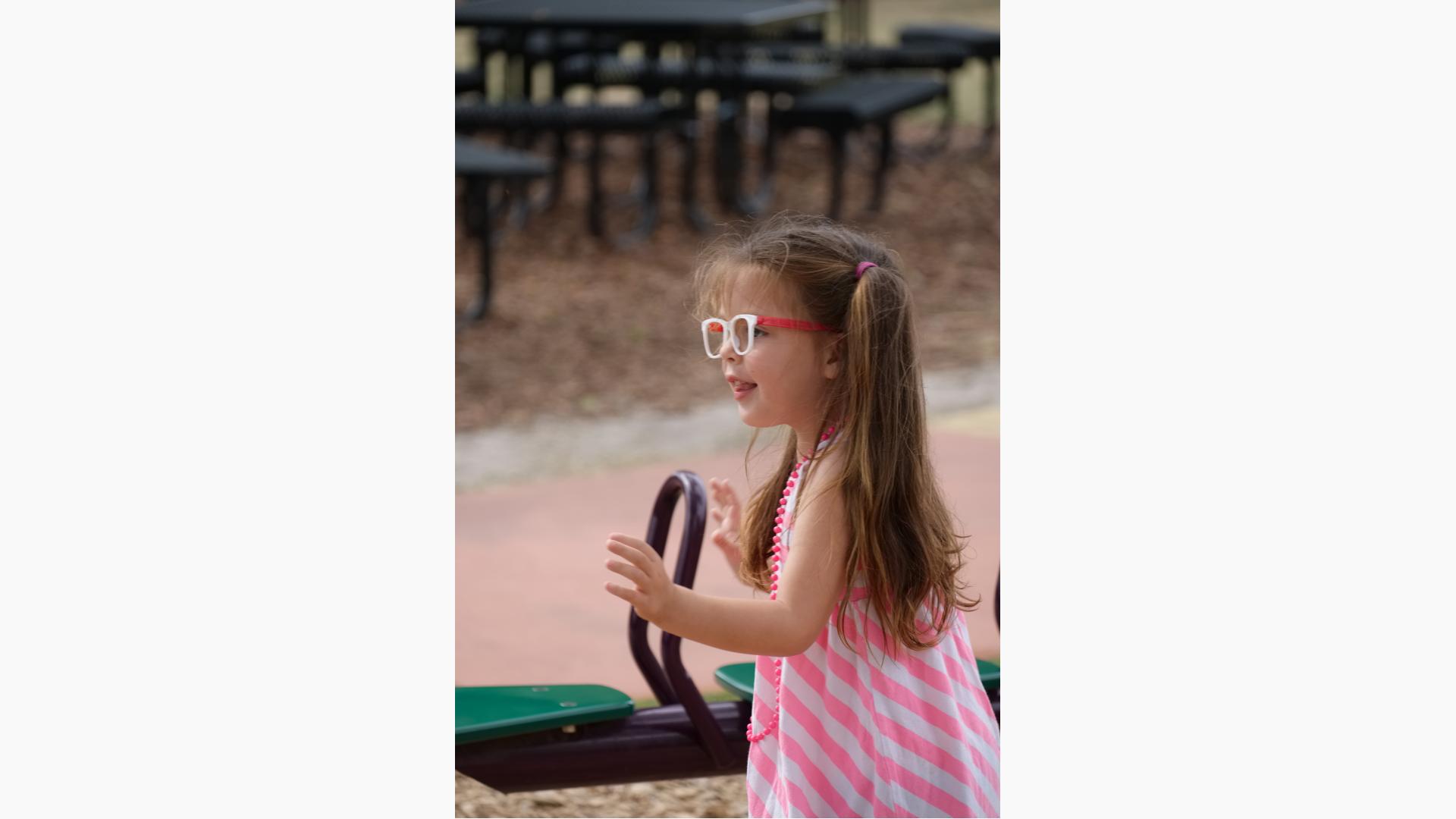 Girl wearing pink stripe dress and glasses stands next to see-saw