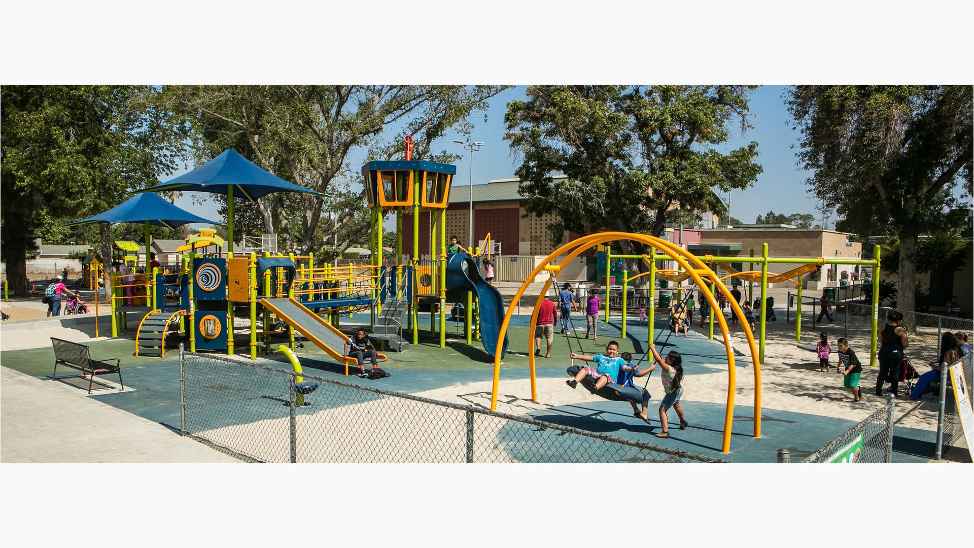 Children in Sepulveda Recreation Center playground. Three kids play on the Oodle swing. A young boy rides down the rollerslide. Parents walk through the playground.