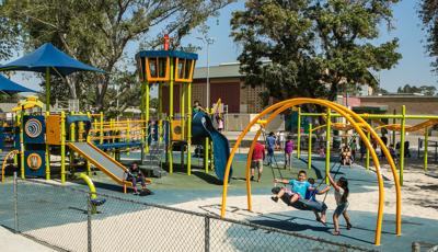 Children in Sepulveda Recreation Center playground. Three kids play on the Oodle swing. A young boy rides down the rollerslide. Parents walk through the playground.