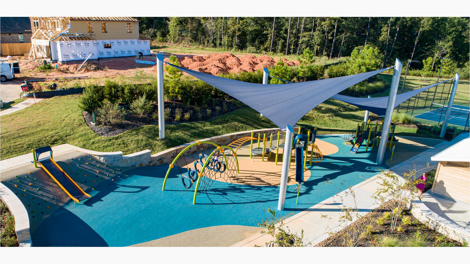 Large dark blue triangular shade sails stand over two separate play structures of a play area. In the background is a construction site of neighborhood homes.
