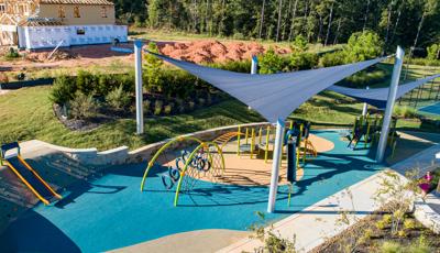 Large dark blue triangular shade sails stand over two separate play structures of a play area. In the background is a construction site of neighborhood homes.