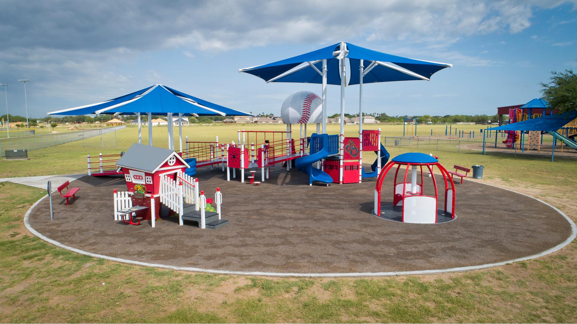 A designated circular play area has one large inclusive baseball themed play structure with accessible ramps, play panels, and two large hexagonal shade systems overhead. A second small play structure designed like a small home sits in front of the large playground along with a inclusive manual carousel. 
