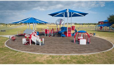 A designated circular play area has one large inclusive baseball themed play structure with accessible ramps, play panels, and two large hexagonal shade systems overhead. A second small play structure designed like a small home sits in front of the large playground along with a inclusive manual carousel. 