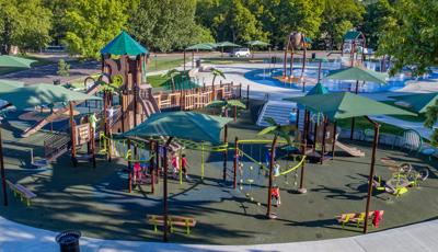Families play at a large play space on a jungle themed playground with  brown posts and green accents and matching near by splash pad. 