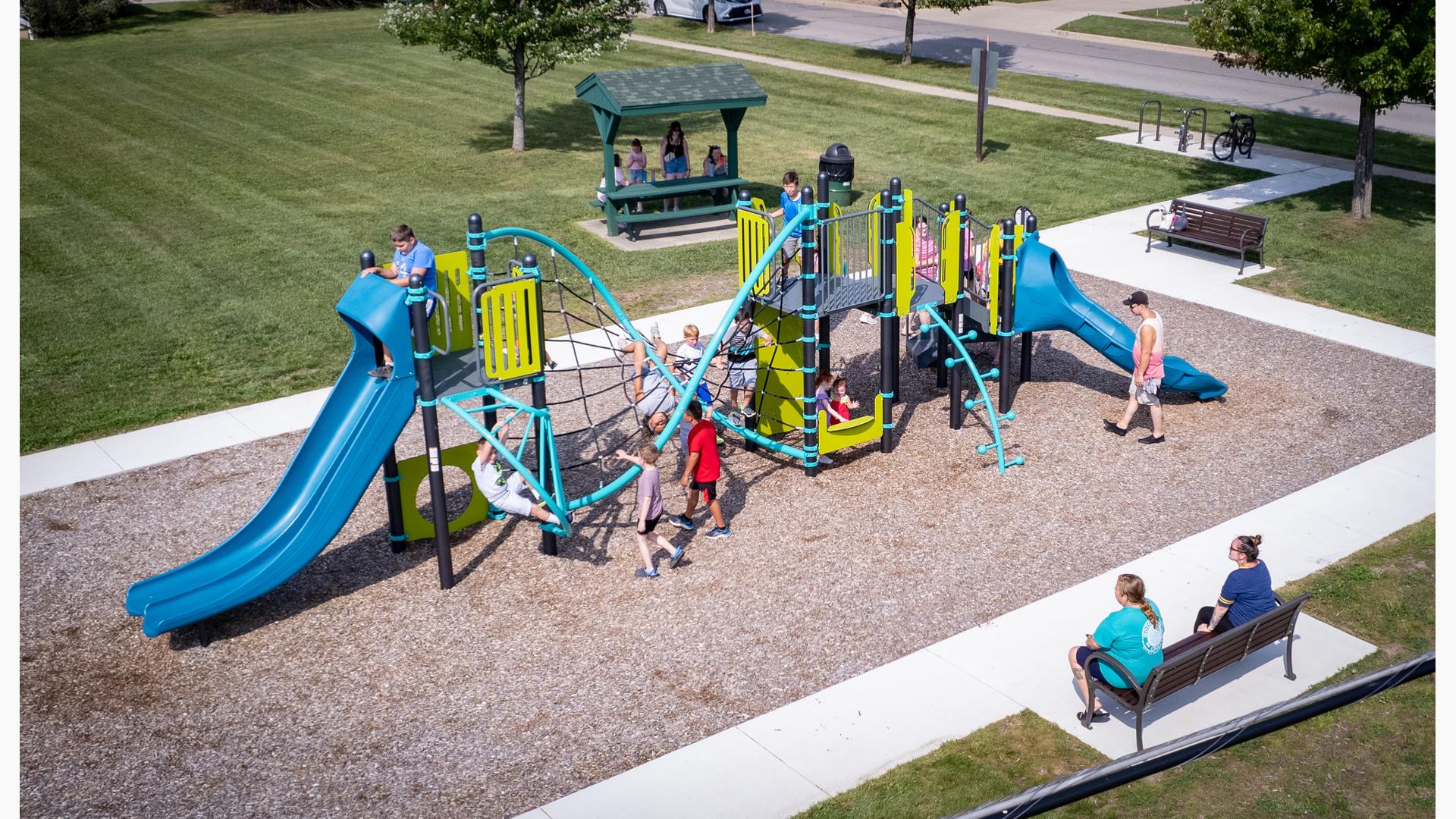 Elevated view of a park playground filled with playing children as parents sit on a bench on the surrounding concrete sidewalk. 
