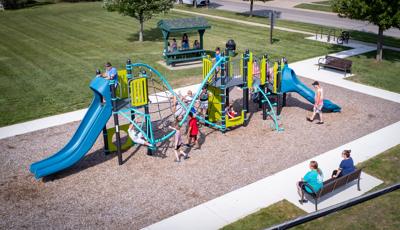 Elevated view of a park playground filled with playing children as parents sit on a bench on the surrounding concrete sidewalk. 