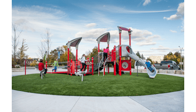Sitting on a green, circular piece of grass, a boy rides down the slide while two girls running up the access ramp. Boy in red shirt sitting on Saddle Spinner.