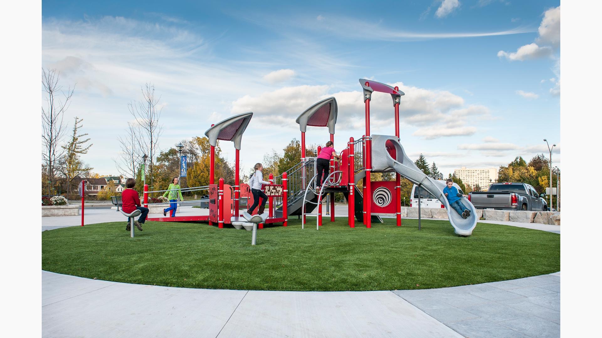 Sitting on a green, circular piece of grass, a boy rides down the slide while two girls running up the access ramp. Boy in red shirt sitting on Saddle Spinner.