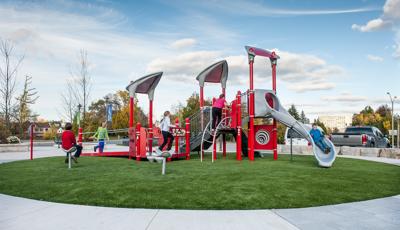 Sitting on a green, circular piece of grass, a boy rides down the slide while two girls running up the access ramp. Boy in red shirt sitting on Saddle Spinner.