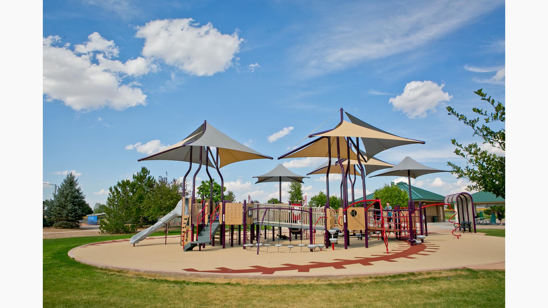 Sports-themed playground with grey and brown multi-panel shade covering the playground.  Surfacing is designed like a baseball. 