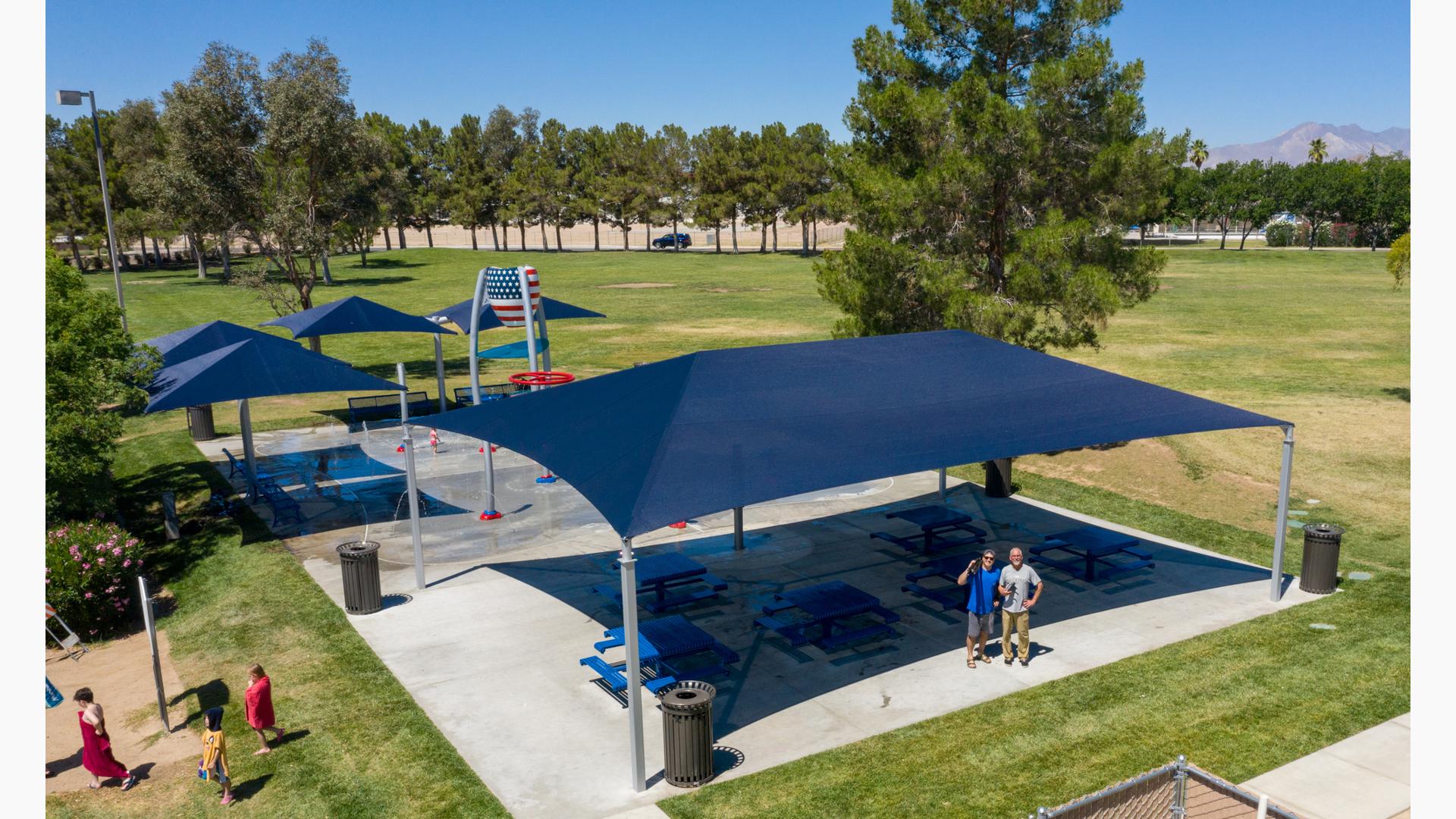 Several navy blue shade structures cover picnic tables and also benches at a splash pad that includes an American flag painted on a dumping bucket along with ground sprays. 