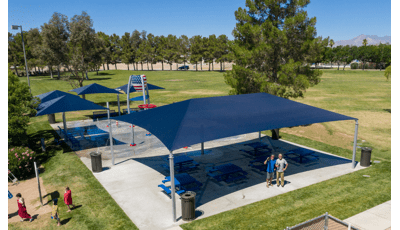 Several navy blue shade structures cover picnic tables and also benches at a splash pad that includes an American flag painted on a dumping bucket along with ground sprays. 