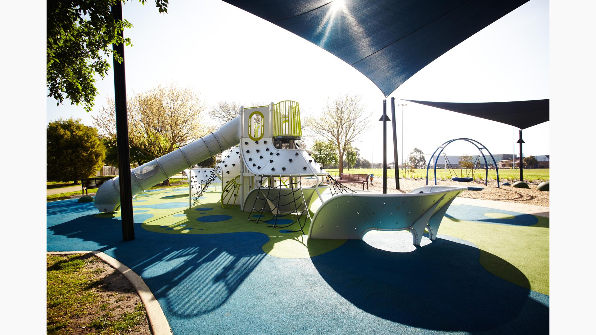 Metal Futuristic playground with multiple panel and net climbers and high, long slide. Blue Shade covering playground and swing in background. Blue and green surfacing with trees on a sunny sky.