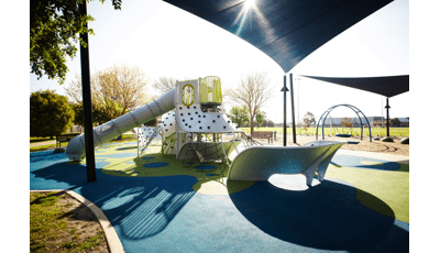 Metal Futuristic playground with multiple panel and net climbers and high, long slide. Blue Shade covering playground and swing in background. Blue and green surfacing with trees on a sunny sky.