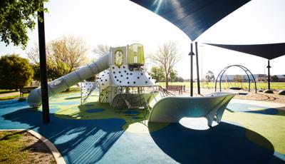 Metal Futuristic playground with multiple panel and net climbers and high, long slide. Blue Shade covering playground and swing in background. Blue and green surfacing with trees on a sunny sky.