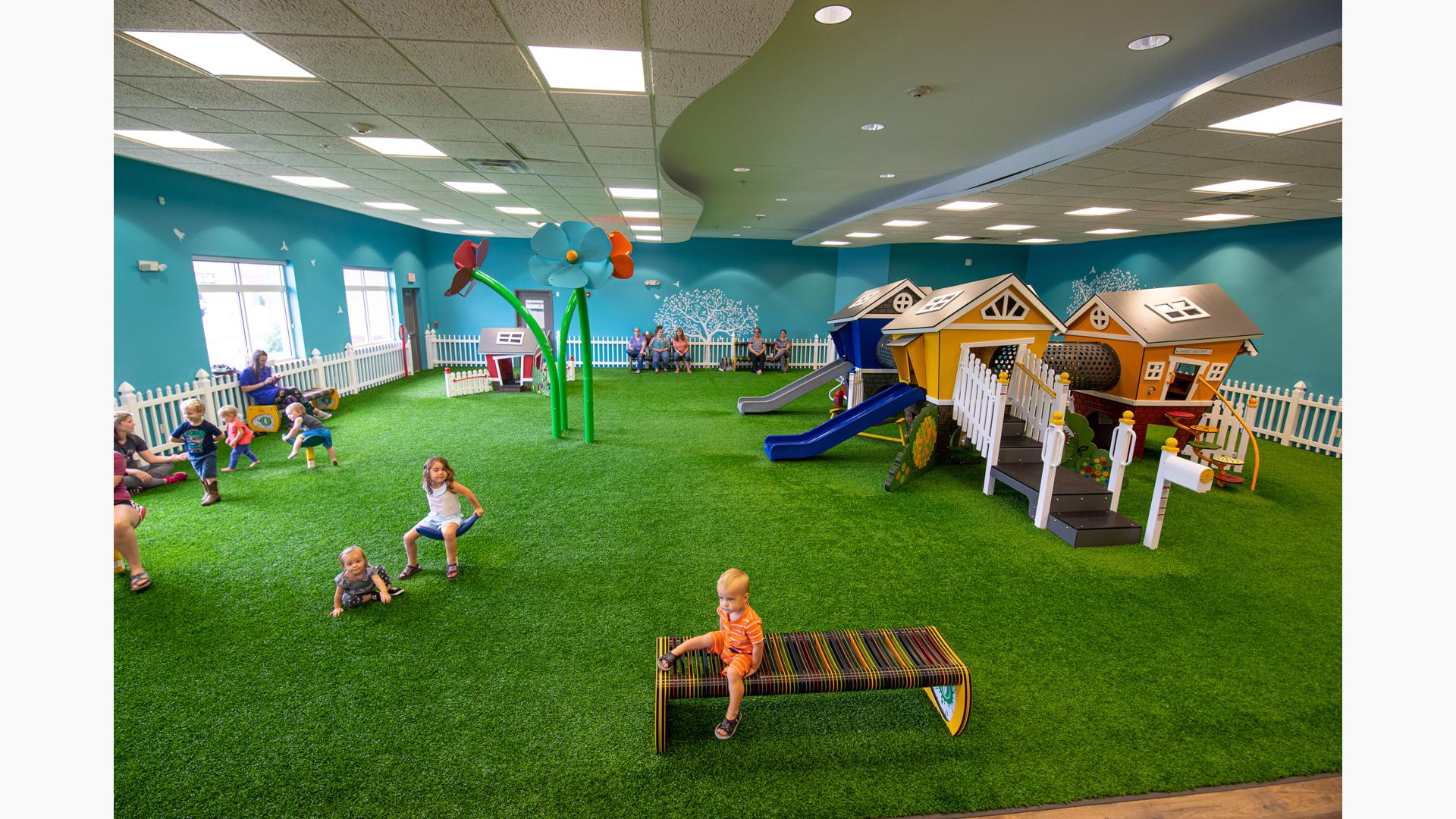 Children play at an indoor play area with artificial grass surfacing. Play structures are designed like little houses accessible for small children.