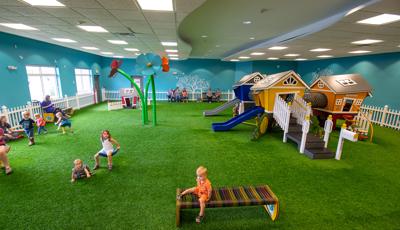 Children play at an indoor play area with artificial grass surfacing. Play structures are designed like little houses accessible for small children.