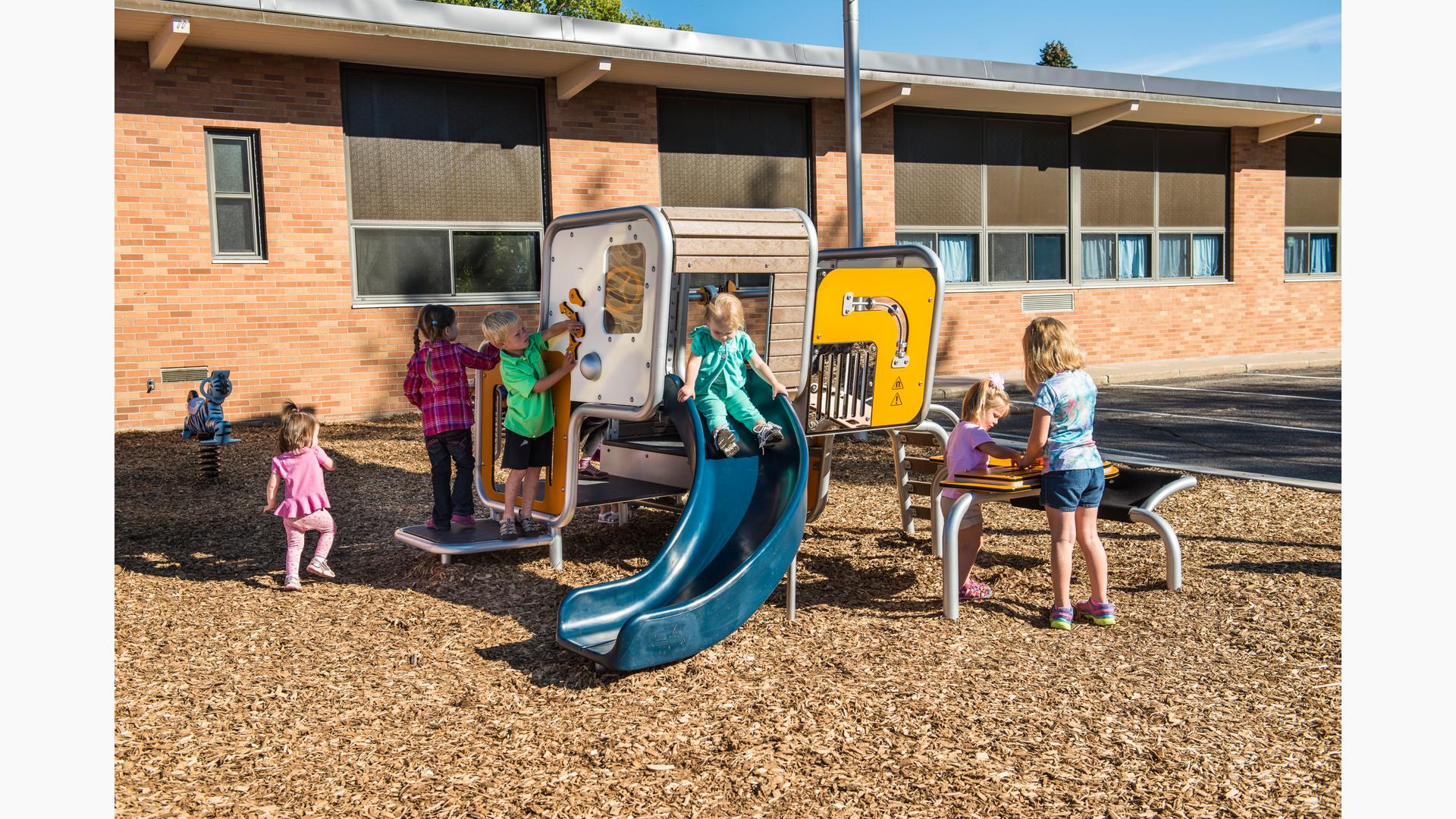 Girl in green outfit sits on top of the Smart Play Cube slide, while a boy in a green shirt plays with the one of the enclosure panels. Two girls play on Grip Desk.