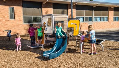 Girl in green outfit sits on top of the Smart Play Cube slide, while a boy in a green shirt plays with the one of the enclosure panels. Two girls play on Grip Desk.