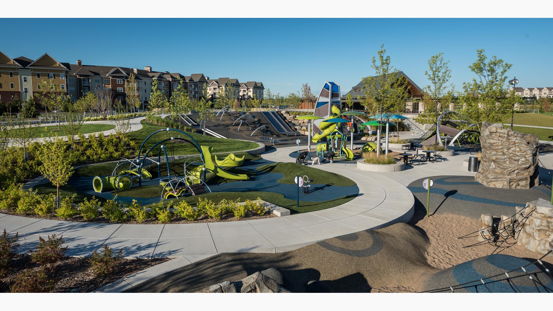 Park with several play areas including a rock climber, enclosed playground tower, climbing net climber and hillside slides with trees intermixed among the play spaces. 
