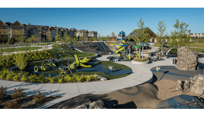 Park with several play areas including a rock climber, enclosed playground tower, climbing net climber and hillside slides with trees intermixed among the play spaces. 