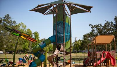 A tall playground tower with a large shade has a spiral belt climber up to the enclosed play area with two large spiral slides. A large flower designed shade with rainbow colored fabric sits to the left of the large tower as a secondary play structure designed like a picnic basket sits to the right.