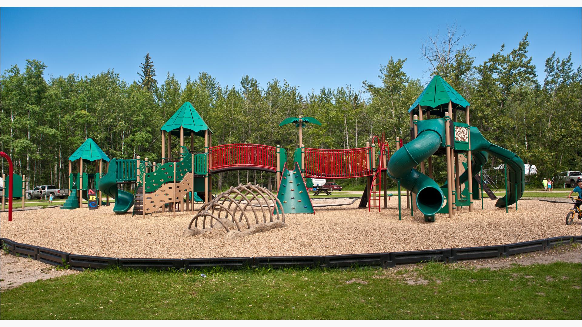 Dinosaur bones penetrate out of the ground in front of a green, red and beige play structure at Pipestone Creek Dinosaur Playground.