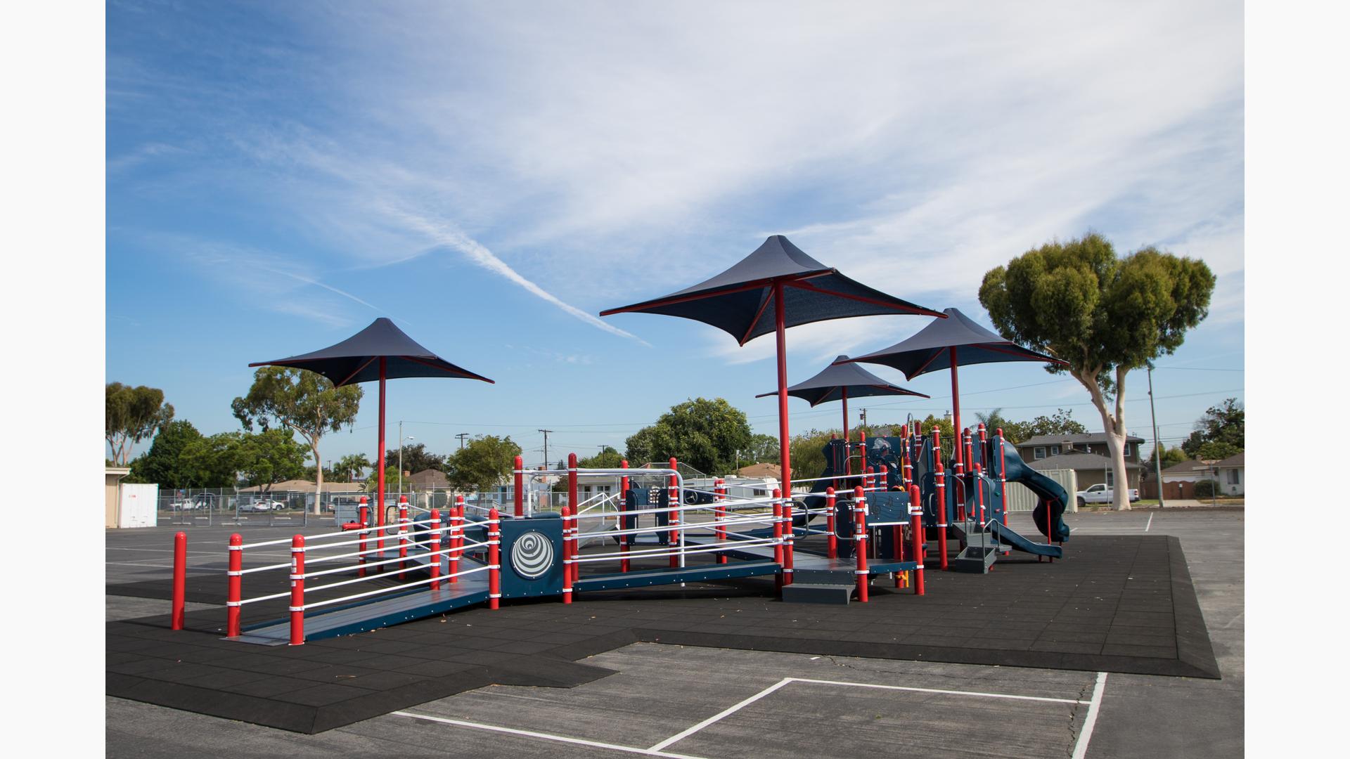 A inclusive school playground with accessible ramps, red posts and navy blue play panels, slides, climbers, and overhead shades.