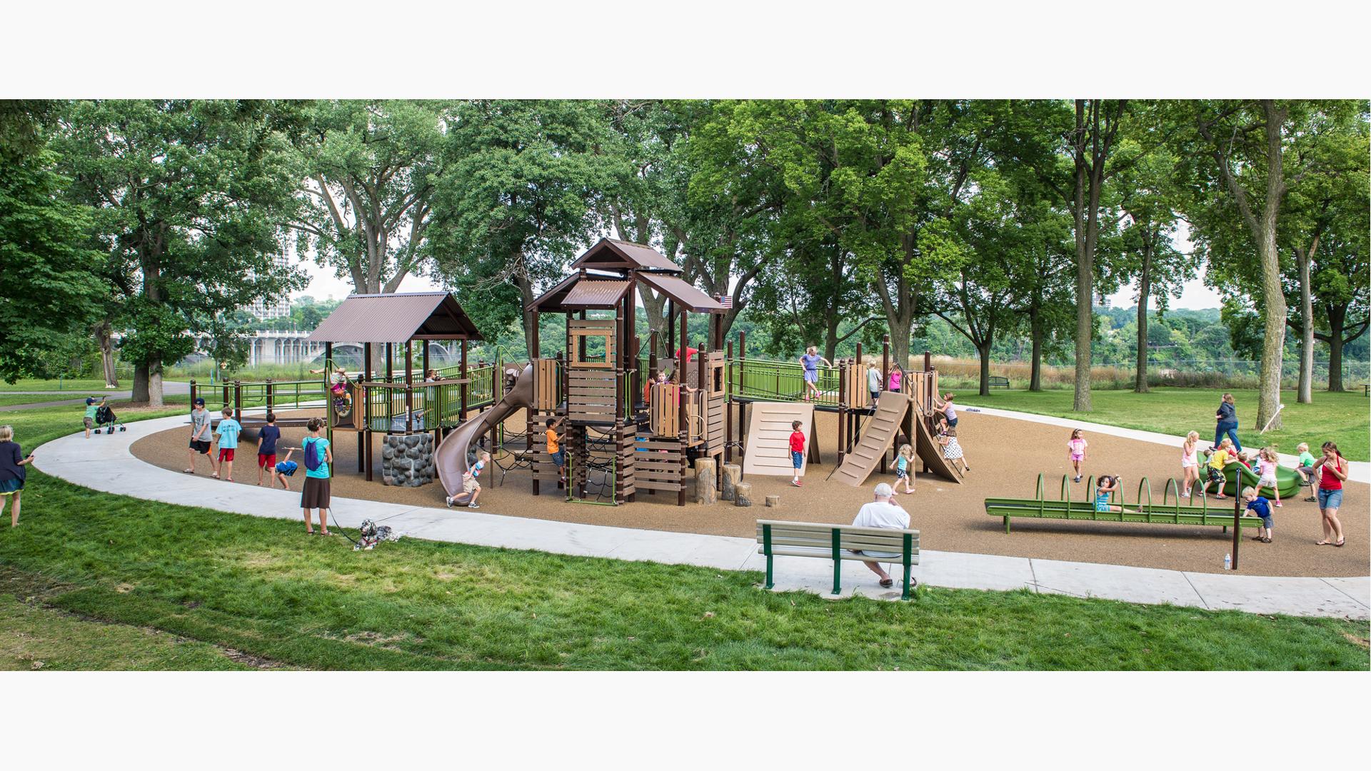 A woman with her dog and an elderly man on a bench watch kids running and playing on the Wabun Picnic Area playground.