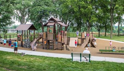 A woman with her dog and an elderly man on a bench watch kids running and playing on the Wabun Picnic Area playground.