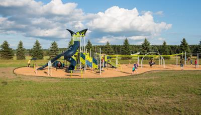 Multiple children playground on playground on a sunny day. Playground includes a tower with two dark blue slides. Play space also includes a slide and zipline with airplane on top. 