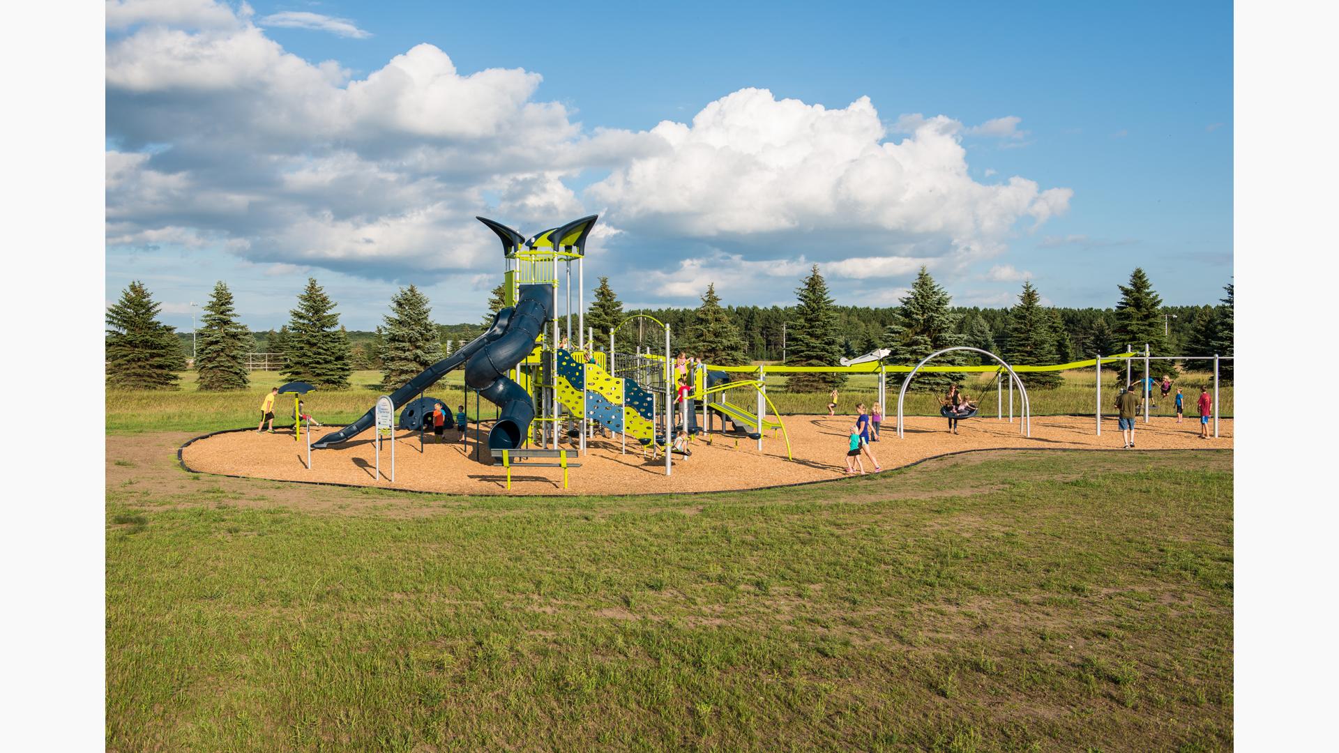 Multiple children playground on playground on a sunny day. Playground includes a tower with two dark blue slides. Play space also includes a slide and zipline with airplane on top. 