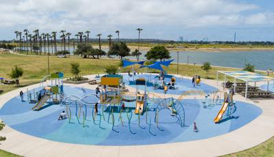 Full elevated view of a large circular play area with multiple play structures and stand alone activities all set next to a large body of water in the background.