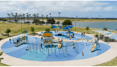 Full elevated view of a large circular play area with multiple play structures and stand alone activities all set next to a large body of water in the background.